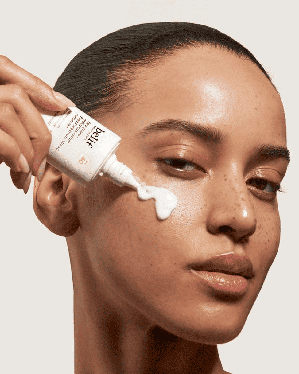 Une main tenant un flacon applicateur dépose une ligne de sérum lacté blanc sur la pommette d'une femme de profil.