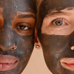 Deux visages, l'un à peau foncée avec un piercing au nez, l'autre à peau claire aux yeux bleus, côte à côte, tous deux couverts d'un masque facial au charbon noir.