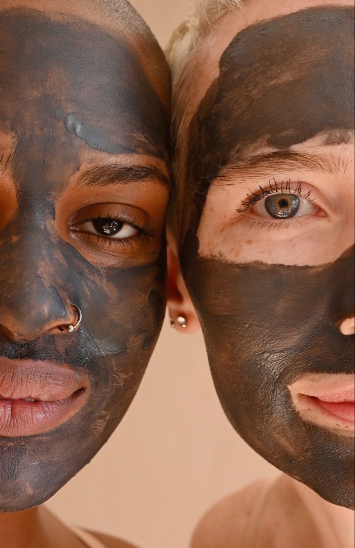 Deux visages, l'un à peau foncée avec un piercing au nez, l'autre à peau claire aux yeux bleus, côte à côte, tous deux couverts d'un masque facial au charbon noir.